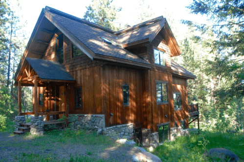 A wooden cabin surrounded by trees, featuring a sloped roof and large windows, set in a natural landscape.